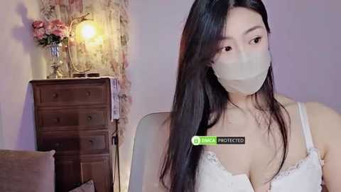 Media: Video of an Asian woman with long black hair, wearing a white lace bra and face mask, standing indoors near a wooden dresser with a lamp and floral decor.