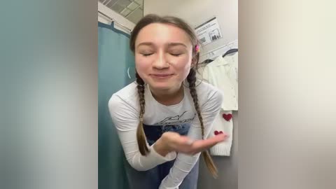 Media: Video of a young Caucasian girl with long braided hair, wearing a white long-sleeve shirt and blue jeans, blowing a kiss, in a hallway with a teal door and white coat rack.