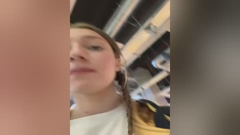 Media: A video showing a young woman with light skin and brown hair, wearing a white shirt and hoop earrings, taken indoors with a blurred background of shelves.