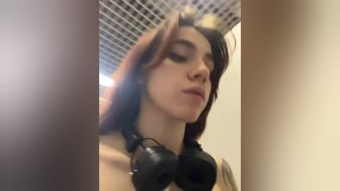 Media: A video of a young woman with long, wavy brown hair, wearing black headphones, looking down. The background shows a grid-patterned ceiling and beige walls. The image has a soft focus.