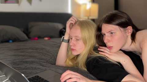 Media: Video of two women in bed, one blonde with a smartwatch, another brunette with short hair, both using laptops; background shows a gray bedspread and a lamp.