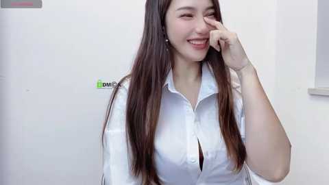 Media: Video of a smiling young Asian woman with long brown hair, wearing a white button-down shirt, and leaning against a white wall, with a window to the right.