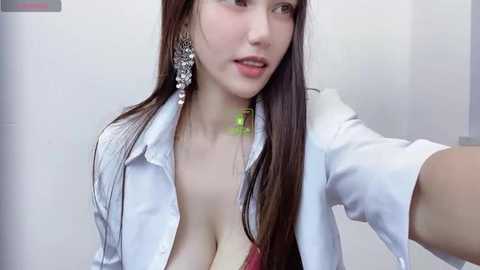 Media: Video of an East Asian woman with long brown hair, fair skin, and large earrings, wearing a white shirt with a plunging neckline, revealing cleavage, taking a selfie in a bathroom.