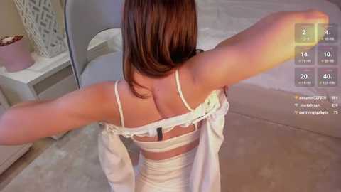 Media: Video of a woman with straight, light brown hair, wearing a white lace bra, pulling a white dress over her head in a bedroom with beige carpet and a white bed.