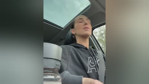 Media: Video of a young, light-skinned, Asian man with short hair, wearing a black hoodie, sitting in a car with a gray seatbelt, eyes closed, holding a white cup, blurry background.