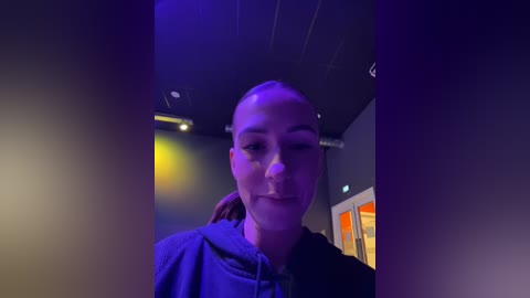 Media: Video of a bald, fair-skinned person with a serene expression, wearing a dark hoodie, standing in a dimly lit room with purple and yellow lighting, and a modern ceiling with recessed lights.