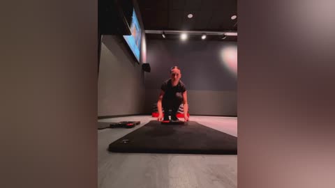 Media: Video of a gym with a woman in a black sports bra and leggings kneeling on a mat, focusing on a large screen above her.