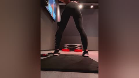 Media: Video of a person in tight black leggings and white sneakers squatting on a black yoga mat in a dimly lit, modern room with gray walls and a flat-screen TV displaying a colorful image.