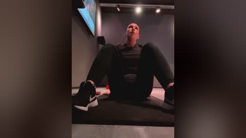 Media: A video captures a man with a muscular physique, wearing a black sports outfit, sitting on a mat in a dimly lit gym. He has a serious expression, with headphones in his ears, and his legs are spread apart.