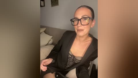 Media: A video of a light-skinned woman with a slim physique, wearing black-framed glasses, a black blazer, and a lace bra, sitting on a beige couch, in a dimly lit room.