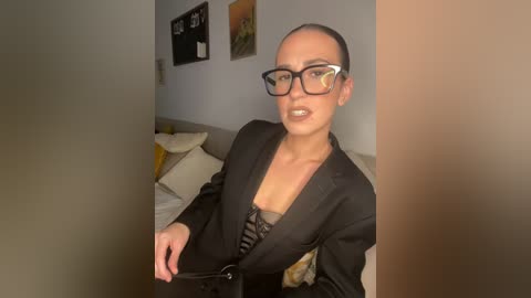 Media: A video of a woman with light skin, black-rimmed glasses, and a black blazer over a floral top, sitting in a modern living room with beige couch and yellow throw pillows.