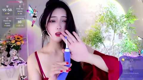 Media: Video of an East Asian woman with pale skin, long black hair, and red lipstick, wearing a red bra, holding a blue vibrator, in a brightly lit room with plants and flowers.