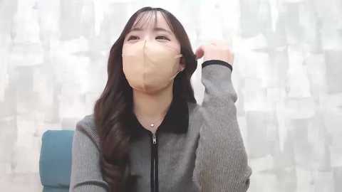 Media: A video of an Asian woman with long, dark hair, wearing a beige face mask and a grey ribbed sweater, adjusting her hair. The background features a light-colored curtain with abstract patterns.