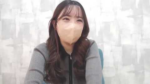 Media: Video of an Asian woman with long dark hair, wearing a beige surgical mask, gray jacket, and backpack, against a white and gray abstract patterned background.