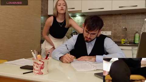 Media: Video of a blonde woman in a black top and white skirt leaning over a man in a blue shirt and vest, writing at a kitchen table.