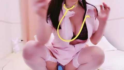 Media: Video of an Asian woman with long black hair wearing a white nurse outfit with a black bra, yellow stethoscope, and white thigh-high stockings, squatting in a white room.