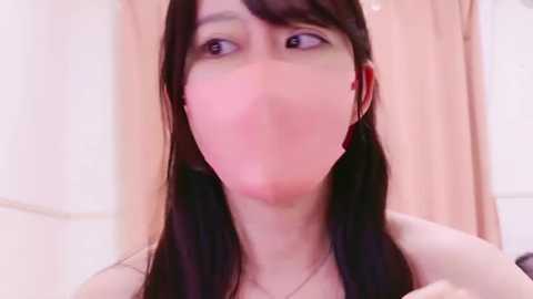 Media: Video of a young Asian woman with long black hair, wearing a beige shirt, and a pink mask, looking slightly distressed. She's in a bathroom with white tiles and a peach-colored curtain in the background.