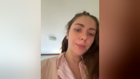 Media: Video of a young woman with fair skin and brown hair, slightly smiling, wearing a pink shirt, partially obscured by a wooden post in a softly lit room with a white wall and a recessed light.