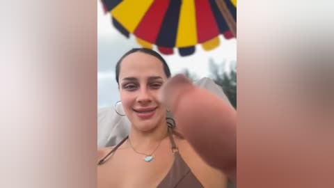 Media: Video of a smiling woman with dark hair, wearing a brown bikini top, holding a large, erect penis in front of her. Background includes a yellow and red umbrella and blurred trees.