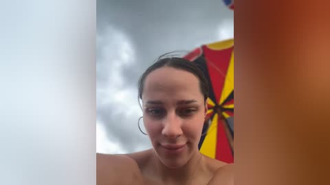 Media: Video of a young woman with dark hair and fair skin, wearing hoop earrings, looking through a hole, partially obscured by a red and yellow umbrella.