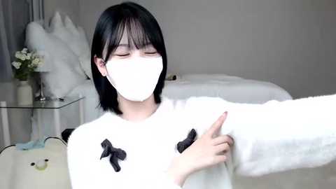 Media: Video of an Asian woman with straight black hair, wearing a white mask, white sweater with black bows, and a white blanket. She's pointing at a stuffed bear. Background features a bed with white sheets and a bedside table with a lamp.