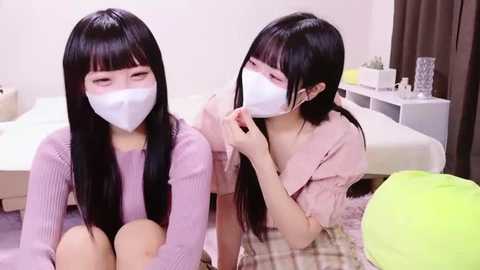 Media: Video of two young East Asian women with long black hair, wearing white masks, light pink sweaters, and sitting on a beige plaid blanket in a cozy, modern room.