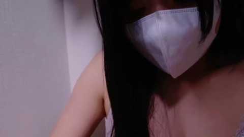 Media: Video of an Asian woman with long black hair, wearing a white surgical mask, revealing her bare shoulders and a hint of cleavage. The background is plain and off-white, creating a minimalistic and clinical atmosphere.