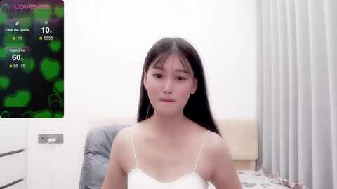 Media: Video of an Asian woman with long black hair in a white lace bra, sitting on a bed in a modern bedroom. A \"Lovense\" screen displaying stats is in the background.