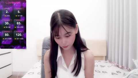 Media: Video of an East Asian woman with long, straight black hair, wearing a white sleeveless top, sitting on a bed with a patterned duvet. The background features a white wall and a purple screen displaying \"Love Live!\" stats.