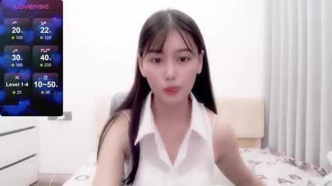 Media: Video of an Asian woman with long black hair and fair skin, wearing a white sleeveless blouse, sitting on a bed with a digital TV displaying a game screen in the background.