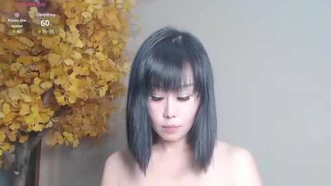 Media: Video of a young Asian woman with straight, shoulder-length black hair, fair skin, and closed eyes, standing against a backdrop of a yellow leafy tree and a muted gray wall.