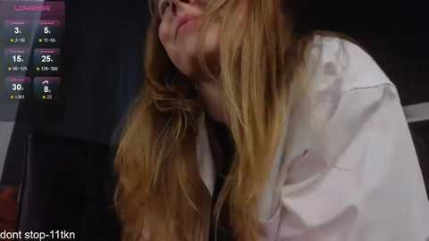 Media: A video of a woman with long, blonde hair partially covering her face, wearing a white shirt, in a dimly lit room.