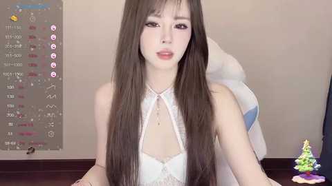 Media: Video of a young East Asian woman with long, straight brown hair, fair skin, wearing a white lace bralette, seated in a chair with a calendar and a small Christmas tree in the background.