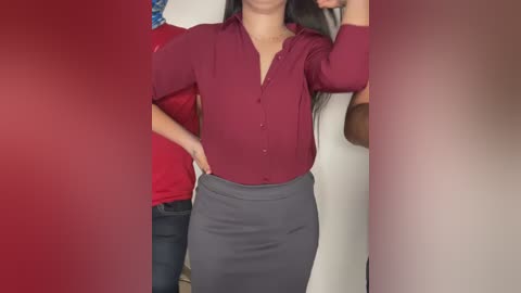 Media: Video of a woman with long black hair, wearing a red blouse and grey pencil skirt, standing in a room with red walls, partially blurred.