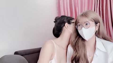 Media: Video of two women in a room with pink curtains; one with glasses and a mask, the other with a white lab coat, kissing passionately.