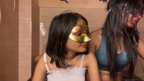 Media: Video of two young Asian women, one in a white tank top, the other in a blue bra, wearing gold Venetian masks indoors.