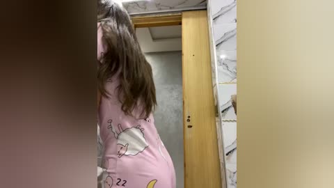 Media: Video of a woman in a pink pajama set with cartoon characters, walking through a modern bathroom with white and beige marble tiles.