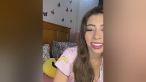 Media: Video of a smiling woman with long brown hair, wearing a pink shirt, in a cozy bedroom with a wooden headboard, butterfly wallpaper, and a plush toy.