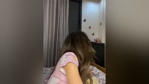 Media: Video of a young woman with long brown hair, wearing a pink pajama top, sitting on a bed with floral sheets, facing a wall decorated with butterfly stickers, in a dimly lit bedroom.