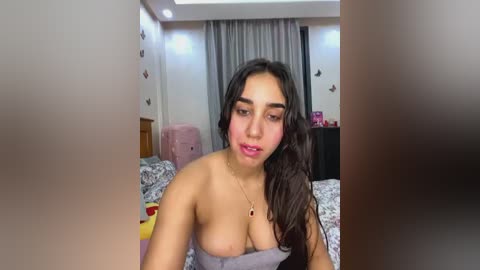 Media: Video of a young woman with long dark hair and light skin, wearing a strapless top, sitting on a bed in a cozy bedroom.