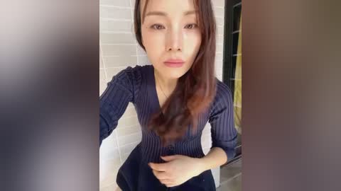 Media: Video of a young Asian woman with straight, dark brown hair, wearing a navy blue, long-sleeve, button-down blouse. She's indoors, standing in front of white tiled walls with a black metal frame in the background.