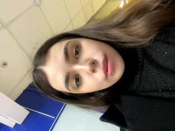 Media: Video of a young woman with long, dark brown hair, light skin, and subtle makeup, wearing a black turtleneck sweater. She stands in a tiled bathroom with blue and white accents, looking directly at the camera.