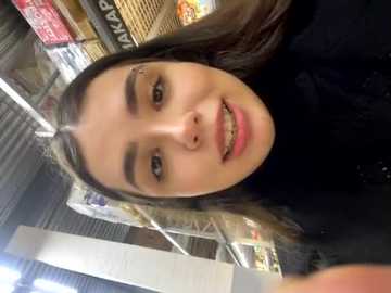 Media: Video of a young woman with straight brown hair, fair skin, and braces, smiling in a supermarket aisle with fluorescent lighting and visible signs in the background.