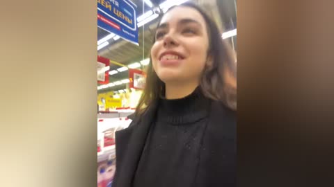 Media: Video of a smiling woman with long brown hair in a grocery store, wearing a black turtleneck sweater, surrounded by shelves filled with various products and a blue and red sign in the background.