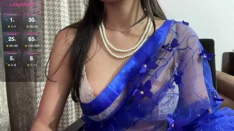 Media: Video of an Indian woman with long black hair, wearing a sheer blue saree with white lace and floral embroidery, revealing a white bra underneath. She's seated, wearing a pearl necklace. Background shows a textured curtain and a weather forecast.