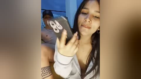 Media: Video of a topless man with tattoos, wearing a cap, and a woman with long dark hair, wearing a low-cut gray hoodie, smiling, holding a smartphone.