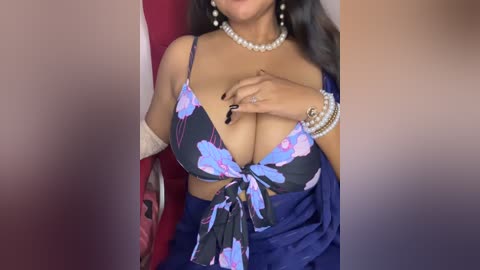 Media: A video of a dark-skinned woman with long black hair, wearing a low-cut, floral-print bikini top and a blue skirt, adorned with pearls and bracelets.