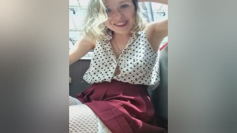 Media: Video of a smiling, fair-skinned woman with blonde, wavy hair, wearing a sleeveless polka-dot blouse, maroon skirt, and white fishnet stockings. She sits in a car, with sunlight streaming through the window behind her.