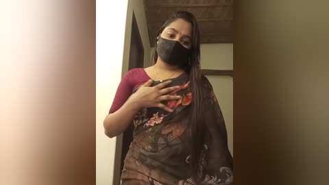 Media: A video of a young woman with long black hair, wearing a black floral sari, a pink top, and a black face mask, standing indoors with wooden beams visible in the background.