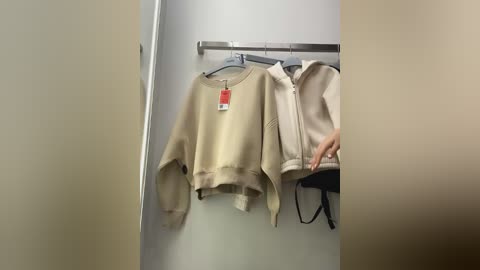 Media: A video of a beige sweater hanging on a metal rack in a beige-walled dressing room. The sweater has a round neckline and long sleeves. To the right, a pair of black undergarments is visible on another rack.